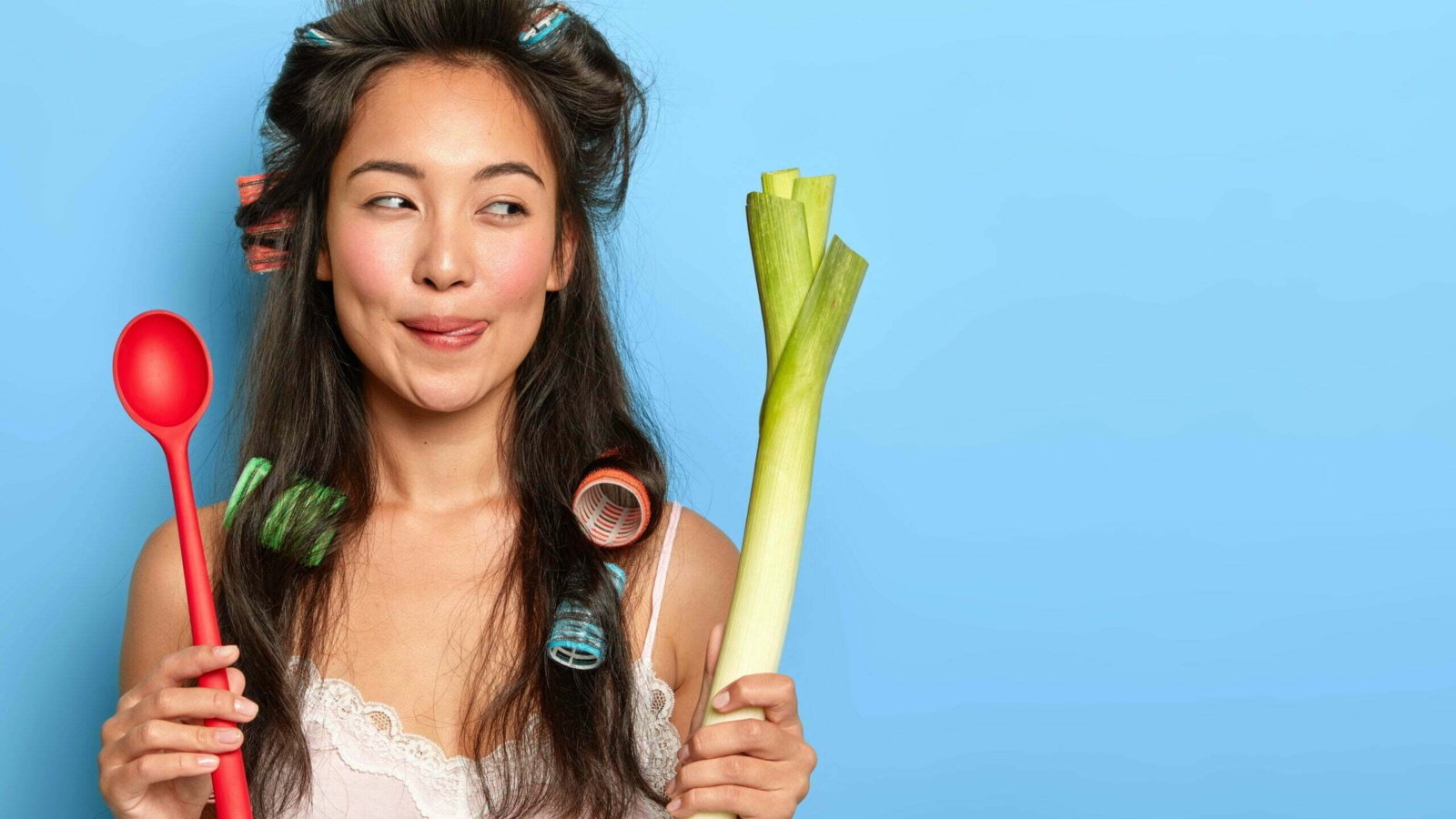Image of pleasant looking brunette housewife with Asian appearance, holds spoon and green leek, prepares vegetarian breakfast, wears curlers for making hairstyle, poses indoor against blue background