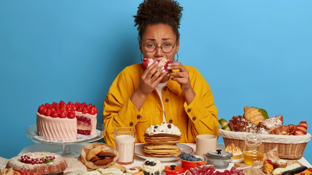 Gluttony and overeating concept. Upset crying ethnic woman eats piece of cake reluctantly, sits at table with many desserts, isolated over blue wall, feels hungry and greedy, wears yellow jacket