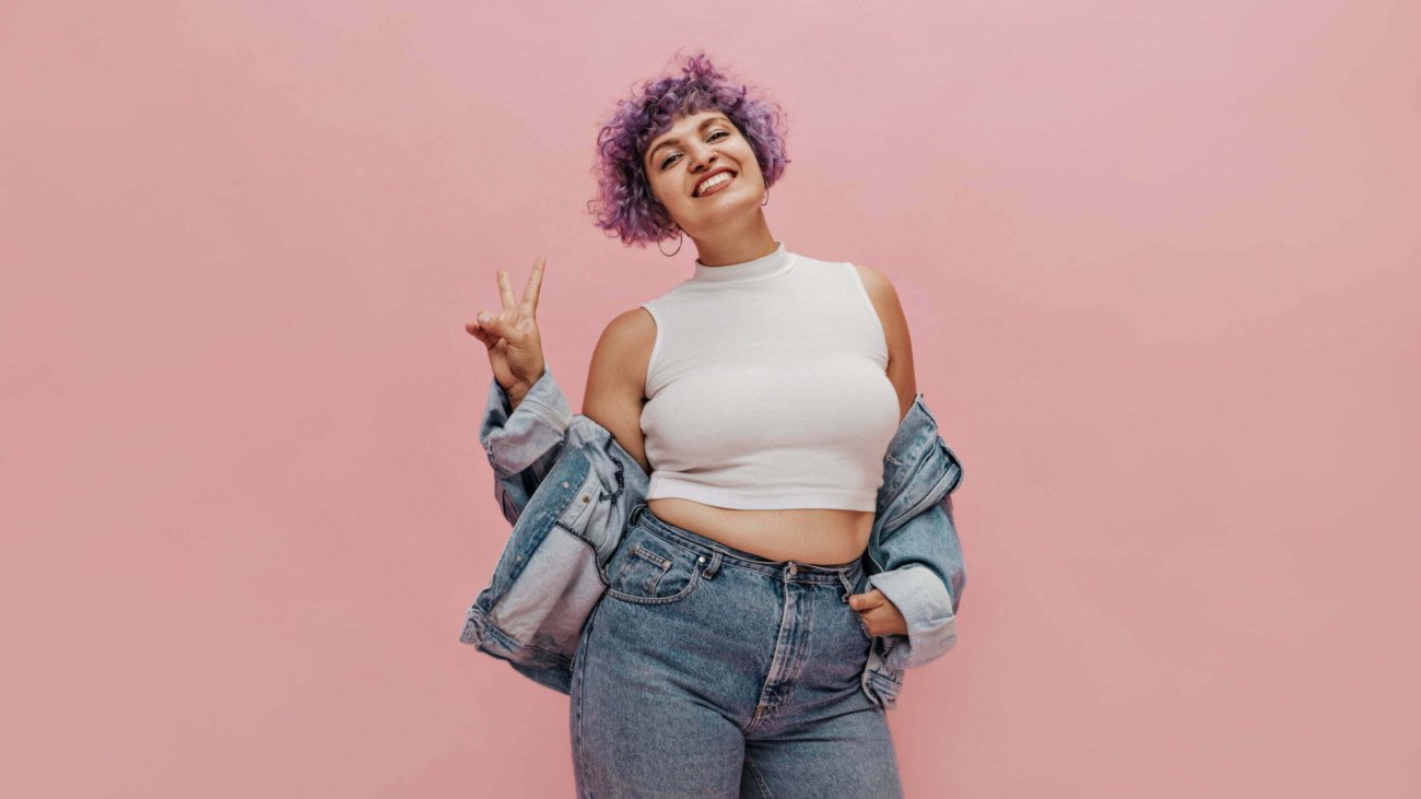 Funny curly girl with lilac hair with lovely smile in white T-shirt posing on camera shows peace signs on pink background..