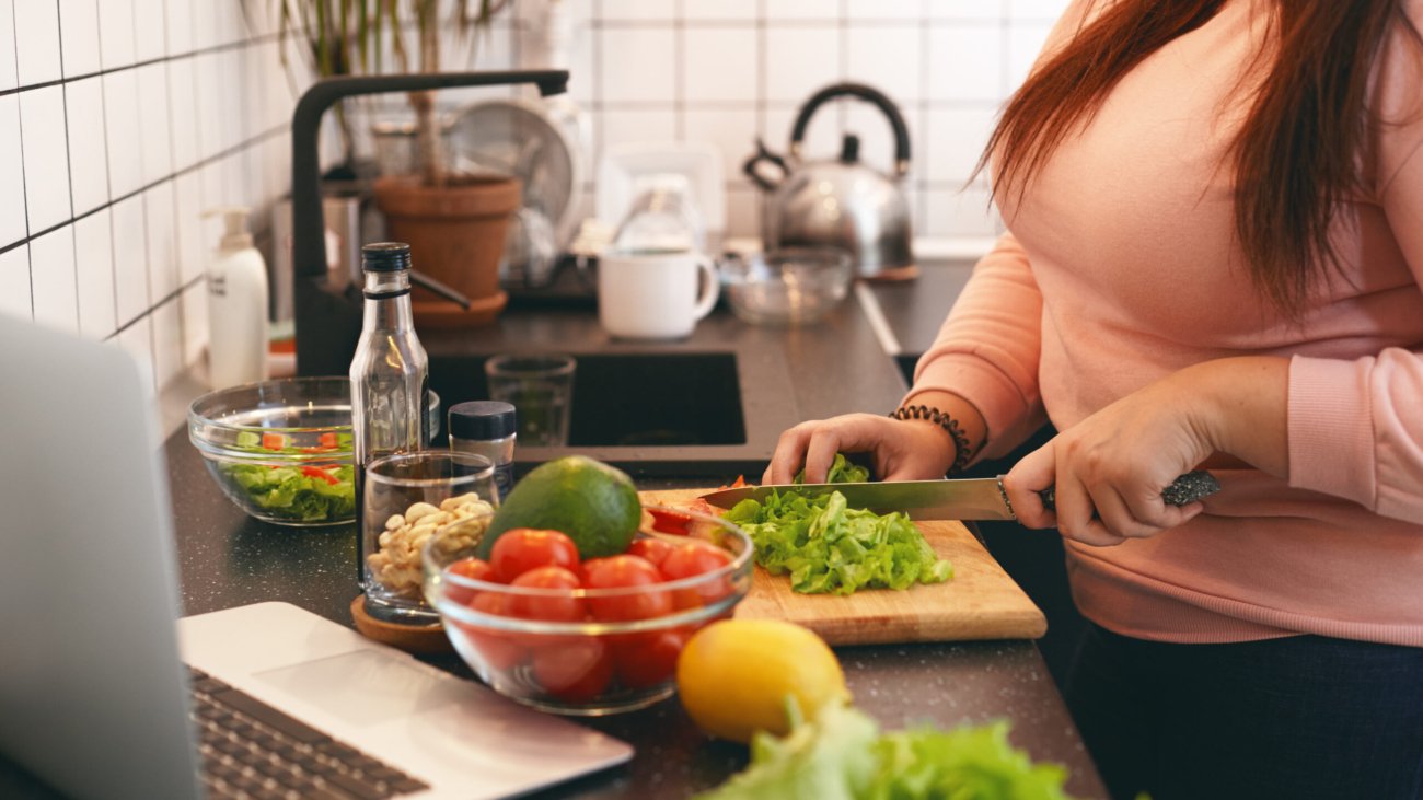 Unrecognizable plus size female standing at kitchen counter and cutting fresh organic vegetables on chopping board, making healthy low calories salad while watching movie online on laptop computer