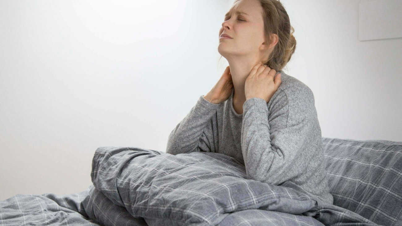 Tired sick young Caucasian woman in grey homewear sitting on bed with closed eyes, touching her neck, suffering from pain. Illness, pain concept