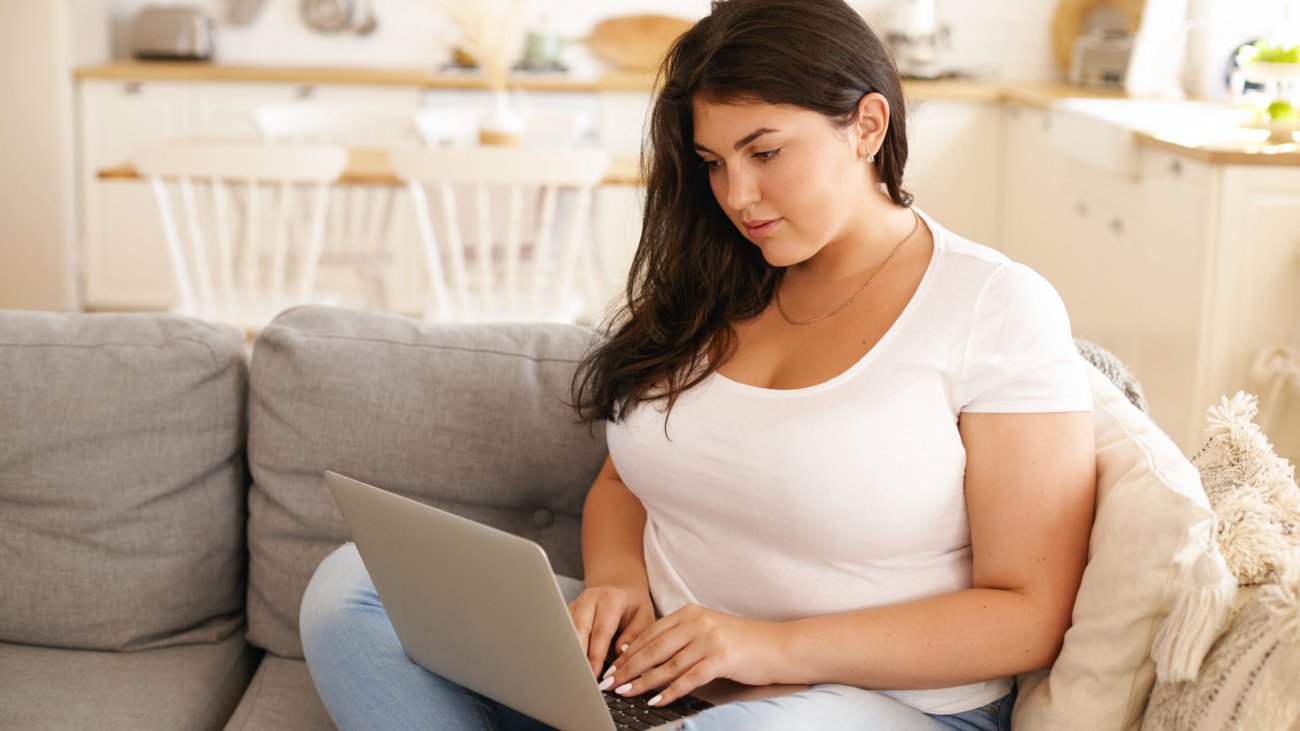 Adorable plus size student girl with loose black hair keyboarding on laptop sitting comfortably on sofa in living room, smiling, messaging friends online, using wireless high speed internet connection