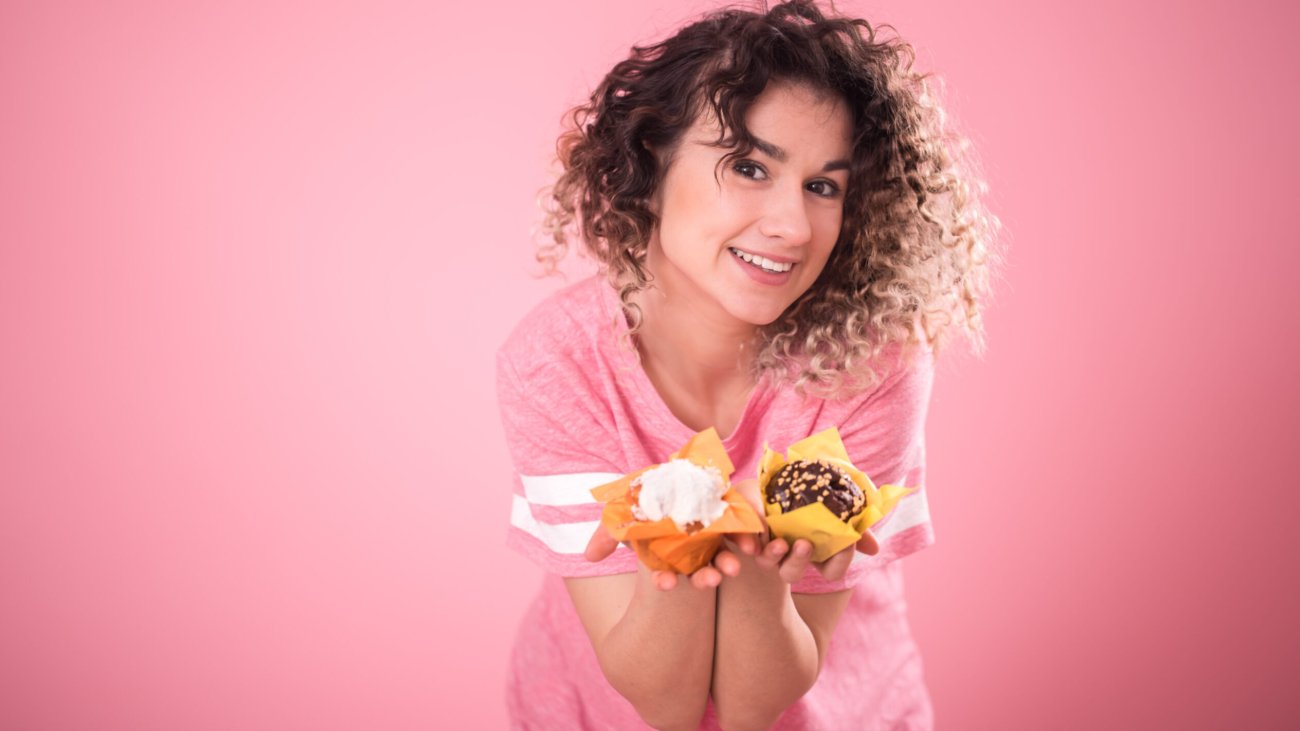 Portrait of a cheerful young curly-haired girl with cupcakes in her hands, in a loose pink t-shirt isolated on a pink background, a place for text, the concept of emotions and healthy eating