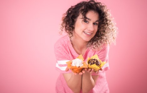 Portrait of a cheerful young curly-haired girl with cupcakes in her hands, in a loose pink t-shirt isolated on a pink background, a place for text, the concept of emotions and healthy eating